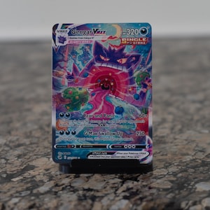 May include: A Gengar VMAX Pokémon trading card. The card features a purple Gengar character with a red and pink energy blast. The card has the text "Fear and Panic" and "G-Max Swallow Up". The card is on a black stand.