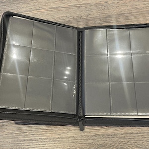 K&ouml;nnte beinhalten: Ge&ouml;ffneter schwarzer Ordner mit transparenten Kunststoffh&uuml;llen, zur Aufnahme von Karten oder Fotos. Der Ordner ist mit einem Rei&szlig;verschluss verschlossen und liegt offen auf einer Holzoberfl&auml;che.