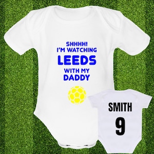Puede incluir: Un body blanco para bebé con texto azul que dice "Shhhh! Estoy viendo Leeds con mi papá" y una pelota de fútbol amarilla. Un body blanco para bebé con texto negro que dice "Smith 9" en la espalda.