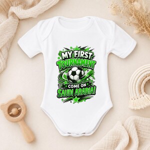 May include: A white short-sleeved baby bodysuit featuring a graphic print of a football surrounded by green and black abstract shapes and stars. The text reads "MY FIRST TOURNAMENT" and "COME ON SAUDI ARABIA!" in bold green and white lettering.