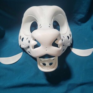 May include: White unfinished furry animal mask with a detailed nose and mouth. The mask is made of a smooth, hard plastic and has a textured surface. The mask is missing the eyes and ears.