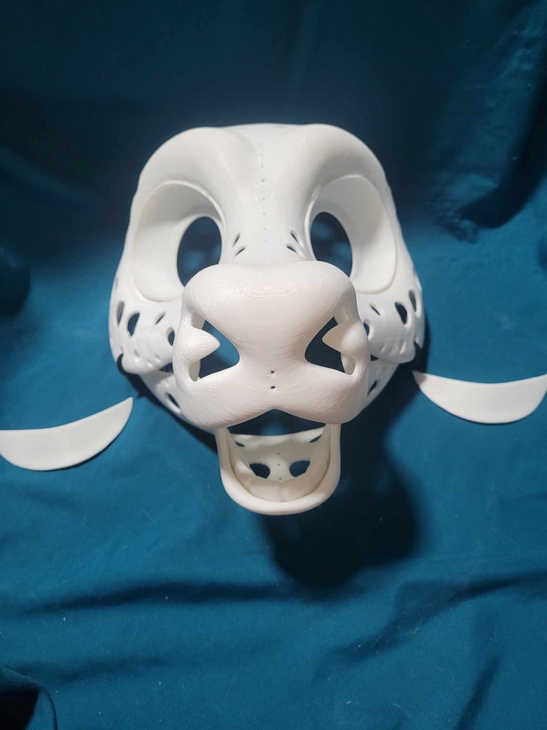 May include: White unfinished furry animal mask with a detailed nose and mouth. The mask is made of a smooth, hard plastic and has a textured surface. The mask is missing the eyes and ears.