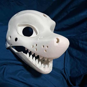 Könnte beinhalten: Eine weiße, 3D-gedruckte Tiermaske mit glatter, matter Oberfläche, großen Augenhöhlen, einer markanten Schnauze mit kleinen Belüftungslöchern und einer Reihe scharfer, spitzer Zähne entlang der offenen Kieferpartie des Kopfes.