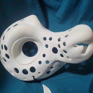 May include: White 3D printed mask with a large hole for the eyes and a smaller hole for the mouth. The mask has a textured surface with many small holes.