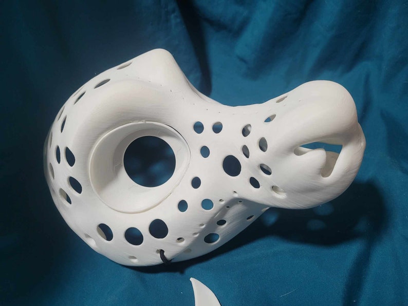 May include: White 3D printed mask with a large hole for the eyes and a smaller hole for the mouth. The mask has a textured surface with many small holes.