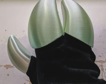 May include: A black glove with three metallic green claw-shaped attachments. The claws have a textured surface and are attached to the glove's fingertips, creating a fantasy or cosplay accessory.