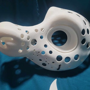 May include: White 3D printed mask with a large hole for the eyes and many smaller holes throughout the mask.