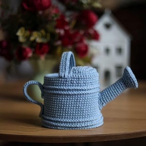 以下が含まれることがあります： 取っ手付きの薄い青色の編み物製のじょうろ。じょうろは木製のテーブルの上に置かれています