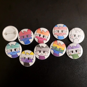 Puede incluir: Un conjunto de botones redondos y coloridos con un diseño de gato de dibujos animados con varios esquemas de color de banderas del orgullo. También se incluye un botón en blanco. Los botones están dispuestos sobre una superficie oscura.
