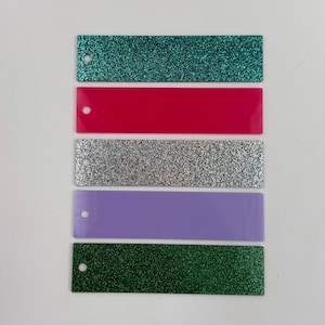 May include: Five different colored glitter bookmarks. The bookmarks are all the same size and shape. The colors are teal, pink, silver, lavender, and green.