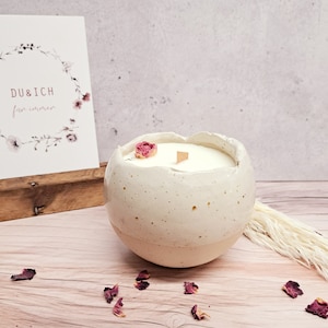 Könnte beinhalten: Ein weißer Kerzenhalter aus Keramik in Form eines Eies mit einer weißen Kerze darin. Der Kerzenhalter hat eine gesprenkelte Oberfläche und eine kleine rosa Blume oben. Die Kerze ist von getrockneten Rosenblättern auf einer hellen Holzoberfläche umgeben.