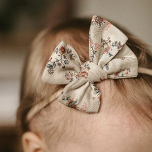 以下が含まれることがあります： クリーム色の背景にピンク、ブルー、グリーンの花が咲いた、花柄のボウが付いたベージュのヘッドバンド。ボウは結び目になっていて、クリーム色の背景にピンク、ブルー、グリーンの花が咲いています。