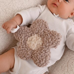 Puede incluir: Un bebé con un body blanco sostiene un juguete de peluche con forma de flor. El juguete es de color marrón claro con un centro color crema. El bebé está acostado sobre una superficie suave y texturizada.