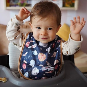 Può includere: Un bambino che indossa un bavaglino blu e marrone con un motivo a tema spaziale. Il bavaglino ha una tasca e un'etichetta bianca con il testo "Love & Ginger".