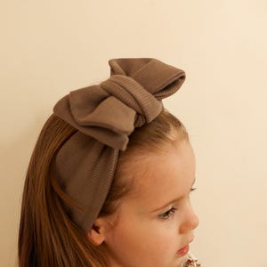 Könnte beinhalten: Ein braunes Stoffhaarband mit einer großen Schleife. Das Haarband wird von einem jungen Mädchen mit langen braunen Haaren getragen.