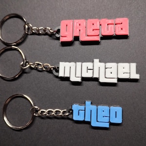Puede incluir: Tres llaveros con diferentes nombres. El llavero superior tiene el nombre "Greta" en letras rosas. El llavero del medio tiene el nombre "Michael" en letras blancas. El llavero inferior tiene el nombre "Theo" en letras azules.