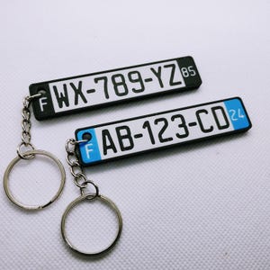 Peut inclure: Deux porte-clés en forme de plaque d'immatriculation noire et blanche avec des anneaux de clés argentés. Le porte-clés du haut porte les lettres "WX-789-YZ" et le numéro "85". Le porte-clés du bas porte les lettres "AB-123-CD" et le numéro "24".