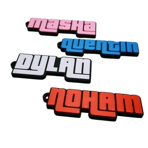 Könnte beinhalten: Vier Namensschilder mit unterschiedlichen Namen: "Masha" in Rosa, "Quentin" in Blau, "Dylan" in Weiß und "Moham" in Rot. Jeder Name ist in einer fetten, stilisierten Schriftart mit schwarzem Rand geschrieben.