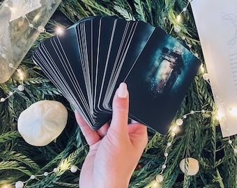 May include: A hand holds a deck of black tarot cards with a single card fanned out, revealing a dark blue and white image with a faint glow. The cards are surrounded by green foliage and white lights.