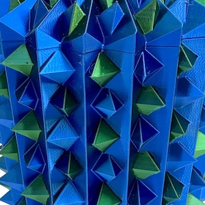 Op de afbeelding: Een geometrische, blauw en groene decoratieve container met een deksel. De container is bedekt met driehoekige vormen, waardoor een getextureerd, stekelig uiterlijk ontstaat. Het deksel heeft een vergelijkbaar ontwerp, met een kleine, diamantvormige bekroning.