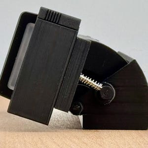 Puede incluir: Un dispositivo mecánico negro con un marco rectangular y una base curva. Una pieza transparente y rectangular es visible dentro del marco. Un resorte dorado está adjunto. El dispositivo descansa sobre una superficie marrón claro.