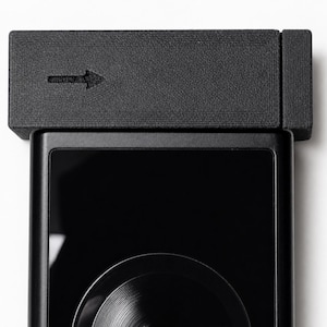 May include: A black electronic device with a textured, rectangular top piece featuring an arrow symbol. The device has a glossy black screen and a circular design element at the bottom. The overall design is modern and minimalist.