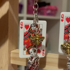 Puede incluir: Pendientes plateados con una carta de la Reina de Corazones, un dije de abeja dorada y un colgante de coraz&oacute;n anat&oacute;mico rojo. Los pendientes cuelgan de una cadena plateada y un llavero.