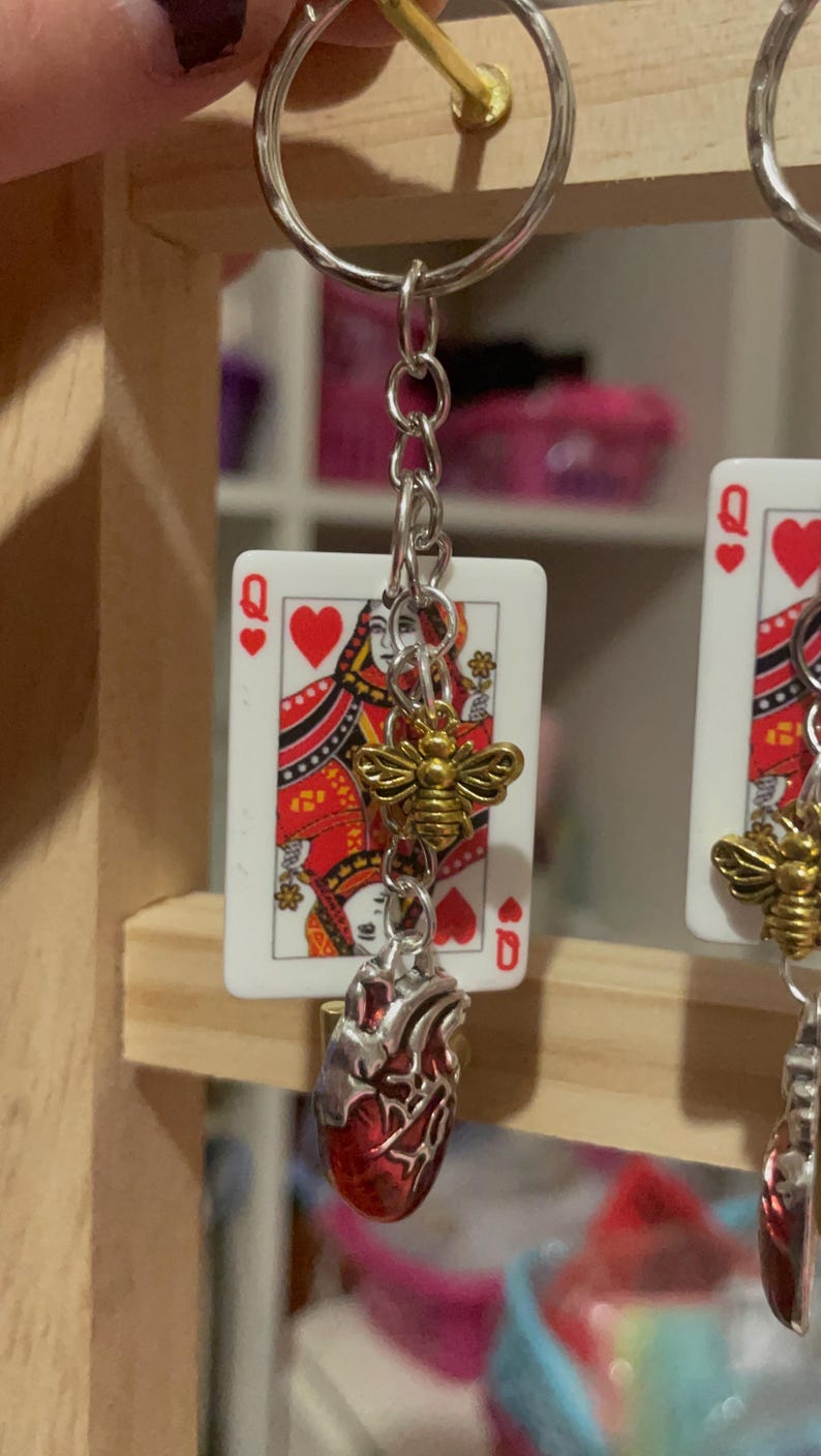 Puede incluir: Pendientes plateados con una carta de la Reina de Corazones, un dije de abeja dorada y un colgante de coraz&oacute;n anat&oacute;mico rojo. Los pendientes cuelgan de una cadena plateada y un llavero.