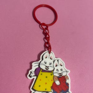 May include: A red keyring with a cartoon image of two white rabbits. One rabbit wears a yellow dress with red dots and a purple top. The other rabbit holds a red stuffed animal and wears a striped shirt.