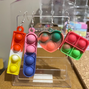 May include: Four pairs of colorful pop it earrings. The earrings are shaped like a heart, a square, and two rectangles. The earrings are made of silicone and have a silver hook.