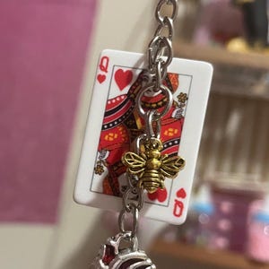 Puede incluir: Un llavero con una carta de la Reina de Corazones, un dije de abeja dorada y un colgante de coraz&oacute;n anat&oacute;mico rojo. El llavero est&aacute; sujeto a una cadena y un anillo plateados.