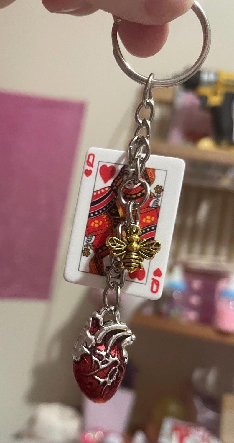 Puede incluir: Un llavero con una carta de la Reina de Corazones, un dije de abeja dorada y un colgante de coraz&oacute;n anat&oacute;mico rojo. El llavero est&aacute; sujeto a una cadena y un anillo plateados.