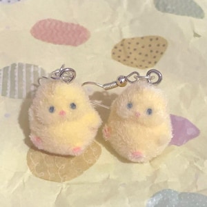 Pode incluir: Um par de brincos ador&aacute;veis ​​de pintinho amarelo. Cada pintinho tem olhos azuis, bico rosa e p&eacute;s rosa. Os brincos s&atilde;o feitos de um material macio e fofo e t&ecirc;m fios de orelha prateados. Em um fundo padronizado.