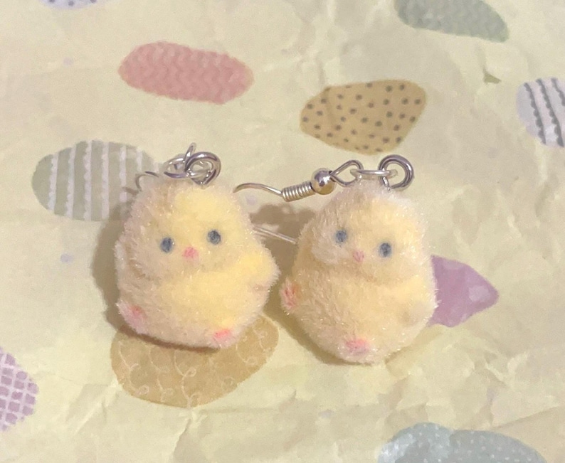 Pode incluir: Um par de brincos ador&aacute;veis ​​de pintinho amarelo. Cada pintinho tem olhos azuis, bico rosa e p&eacute;s rosa. Os brincos s&atilde;o feitos de um material macio e fofo e t&ecirc;m fios de orelha prateados. Em um fundo padronizado.