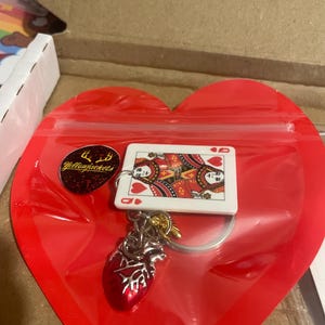 Puede incluir: Un llavero con una carta de la Reina de Corazones y un dije de coraz&oacute;n anat&oacute;mico rojo. El llavero est&aacute; sujeto a un llavero plateado. Los art&iacute;culos est&aacute;n empaquetados en una bolsa de pl&aacute;stico roja en forma de coraz&oacute;n con una pegatina amarilla que dice "Yellowjackets".