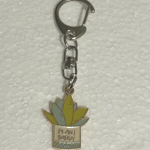 May include: A silver keychain with a metal charm featuring a green and yellow plant in a white pot with the text "Plant Daddy".