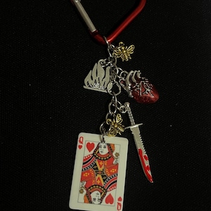 May include: A red heart-shaped carabiner clip with various charms. Charms include a Queen of Hearts playing card, a red heart, a silver knife with red accents, a silver flame, and two gold-coloured bee charms.