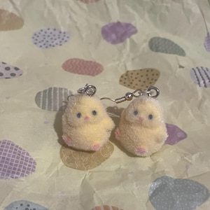 Pode incluir: Um par de brincos de pintinho amarelo com ganchos prateados. Os brincos s&atilde;o fofos e t&ecirc;m p&eacute;s rosa e olhos pretos pequenos. O fundo &eacute; um papel amarelo claro com formas de ovo em cores pastel.