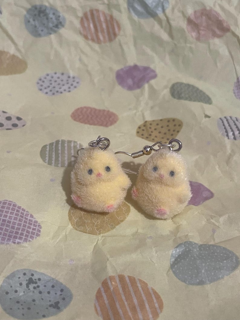 Pode incluir: Um par de brincos de pintinho amarelo com ganchos prateados. Os brincos s&atilde;o fofos e t&ecirc;m p&eacute;s rosa e olhos pretos pequenos. O fundo &eacute; um papel amarelo claro com formas de ovo em cores pastel.