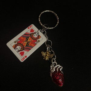 Puede incluir: Un llavero plateado con una carta de la Reina de Corazones, un dije de abeja dorada y un colgante de coraz&oacute;n anat&oacute;mico rojo. El llavero est&aacute; sobre un fondo negro. La carta tiene detalles rojos y dorados.