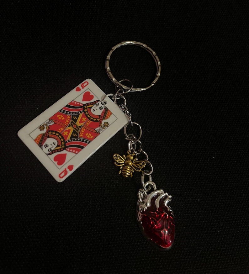 Puede incluir: Un llavero plateado con una carta de la Reina de Corazones, un dije de abeja dorada y un colgante de coraz&oacute;n anat&oacute;mico rojo. El llavero est&aacute; sobre un fondo negro. La carta tiene detalles rojos y dorados.