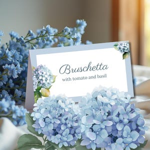 May include: A light blue card with the word "Bruschetta" and the text "with tomato and basil" is surrounded by blue hydrangea flowers and other blue flowers. The card has a light blue border.