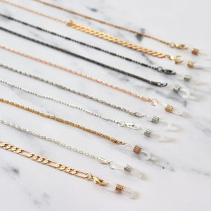 May include: A selection of gold, silver and black eyeglass chains with clear silicone ends. The chains are arranged in a row on a white marble surface.