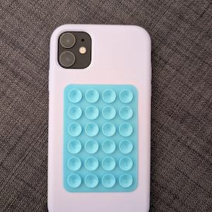 May include: A light purple phone case with a light blue suction cup grip attached. The phone case has a dual-lens camera and flash. The suction cup grip is rectangular with multiple small suction cups.