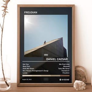 May include: A framed poster featuring the album cover for Freudian by Daniel Caesar. The design includes a central photograph of a person on a slanted roof, the tracklist, the release date August 25, 2017, and a Spotify code on a black background.