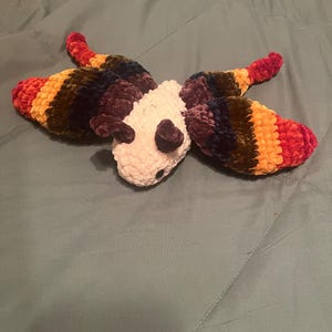 May include: A plush butterfly toy with a white body and multi-colored wings. The wings are striped with red, orange, yellow, green, purple, and blue.