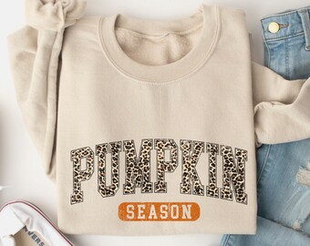 May include: A sand-colored sweatshirt with the text "PUMPKIN SEASON" in a leopard print design. The sweatshirt is paired with light blue jeans and white sneakers. The word "Sand" is at the bottom of the image.