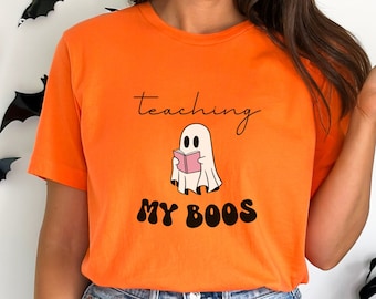 May include: Bright orange t-shirt featuring the phrase "teaching my boos" and a cartoon ghost reading a book. The ghost is white with black eyes and is holding a pink book. The text is in black and brown. The word "Orange" is at the bottom.