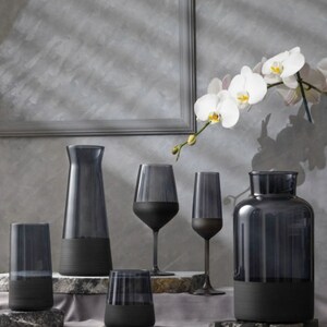 May include: A set of five black glass containers with a frosted finish. The containers include a tall pitcher, two wine glasses, a tumbler, and a vase. The containers are arranged on a gray surface with a white orchid flower in the background.