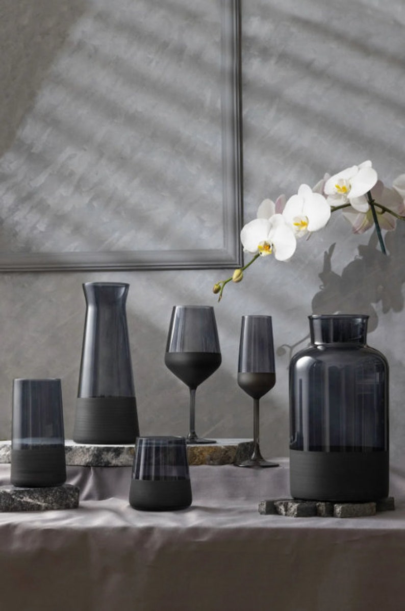 May include: A set of five black glass containers with a frosted finish. The containers include a tall pitcher, two wine glasses, a tumbler, and a vase. The containers are arranged on a gray surface with a white orchid flower in the background.